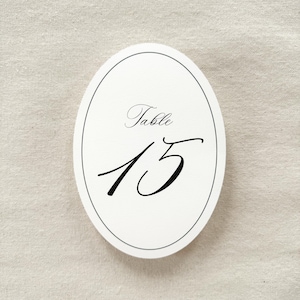 May include: An oval-shaped white table number card featuring a thin black border. The text "Table" is printed in a black script font above the large, prominent black number "15" which is also written in a decorative script typeface.