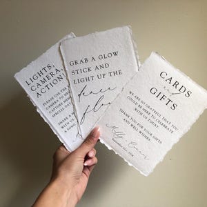 May include: Three white cards with black text. The first card says "Lights, Camera, Action!" and has instructions to use the camera to capture memories. The second card says "Grab a glow stick and light up the dance floor". The third card says "Cards and Gifts" and expresses gratitude for the guests' presence and gifts. The cards are decorated with a rustic, hand-torn edge.