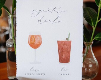 Könnte beinhalten: Eine weiße rechteckige Karte mit Büttenrand zeigt eine Aquarellillustration von zwei Cocktails. Der Text lautet signature drinks, hers Aperol Spritz und his Caesar. Die Karte steht aufrecht auf einem kleinen weißen strukturierten Sockel.