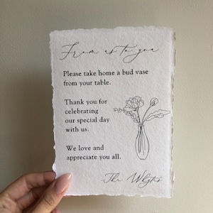 May include: A white card with black text that reads "From us to you. Please take home a bud vase from your table. Thank you for celebrating our special day with us. We love and appreciate you all. - The Wrights." A simple line drawing of a vase with flowers is in the upper right corner.