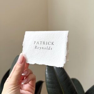 May include: A white, textured paper card with the name "PATRICK Reynolds" printed in black. The card has a deckled edge and is held in front of a plant.