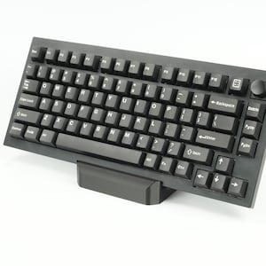Puede incluir: Un teclado mecánico negro con una disposición de teclas negras y blancas. El teclado tiene un reposamuñecas negro y una perilla negra en la esquina superior derecha.