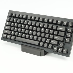 Puede incluir: Un teclado mecánico negro con una disposición de teclas negras y blancas. El teclado tiene un reposamuñecas negro y una perilla negra en la esquina superior derecha.