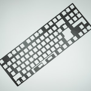 May include: Black metal keyboard plate with cutouts for key switches. The plate is designed for a full-sized keyboard layout.