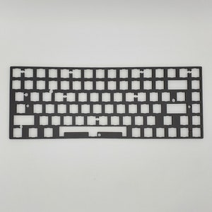 以下が含まれることがあります： キー スイッチ用の切り抜きが施された黒いキーボードプレート。プレートは金属製で、長方形です。
