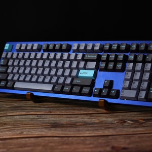 以下が含まれることがあります： 青と黒のメカニカルキーボードで、ライトブルーのキーキャップに「Enter」と表示されています。キーボードは木製の表面に置かれています。