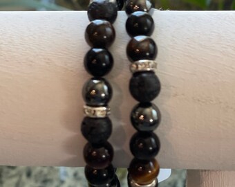 May include: Two beaded bracelets with a variety of dark-colored stones. The bracelets feature black, brown, and tiger's eye beads, interspersed with silver-colored spacers. The bracelets are displayed on a white surface.