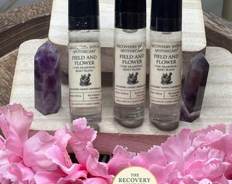 May include: Three glass roll-on bottles with black caps, labeled "Field and Flower The Seasonal Reset Blend" are displayed. The bottles are flanked by purple amethyst crystals and surrounded by pink carnations. The Recovery Witch Apothecary logo is visible.
