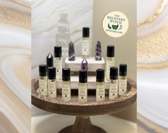 May include: A display of small, clear glass roll-on bottles with black caps, arranged on a wooden stand. The bottles are labeled with text and are surrounded by amethyst and clear quartz crystals. The logo for "The Recovery Witch Apothecary" is visible.