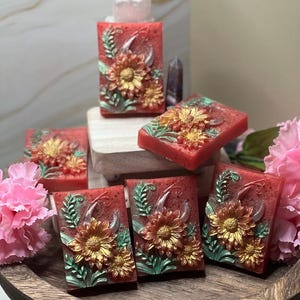 Könnte beinhalten: Rote, rechteckige Seifen mit einem Sonnenblumenmuster in Gold und Grün. Die Seifen sind auf einem Holztablett mit rosa Blumen angeordnet. Ein klarer Kristall und ein violetter Kristall befinden sich im Hintergrund.