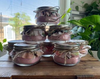 May include: A collection of small, clear glass jars filled with pink wax candles. Each jar has a silver lid, a fabric and twine embellishment. The candles are arranged on a wooden surface, with a window and greenery in the background.
