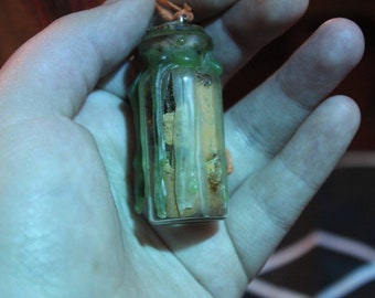 May include: A small, clear glass bottle with a cork stopper, filled with dried plant matter. The bottle is sealed with green wax, and a small orange string is attached to the top. The bottle is held in a hand.