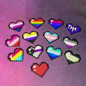 May include: A selection of pixelated heart designs, each with a unique colour scheme. The hearts represent various LGBTQ+ pride flags, including rainbow, transgender, bisexual, and non-binary flags. The background is a soft, blurred purple and pink.