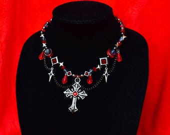May include: A necklace featuring a silver-toned cross pendant with a central red stone, accented by black chains, red teardrop beads, small silver star charms, and round red and grey beads arranged on a silver-toned wire chain structure.