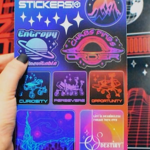 May include: A sheet of space-themed stickers with various designs. The stickers feature illustrations of planets, rovers, and space-related text, including "Entropy," "Chaos Finds Order," and "Destiny." The colour palette includes blues, purples, and oranges.