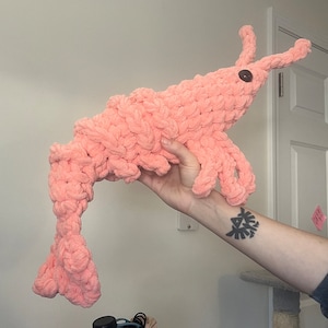 May include: A pink crocheted shrimp plush toy with a brown button eye. The toy is being held by a person with a tattoo on their arm.