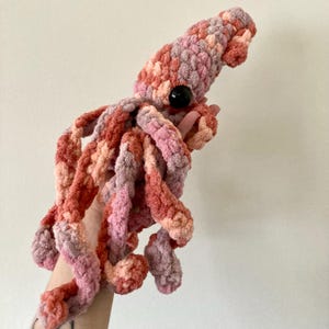 May include: A handmade plush squid in shades of pink, peach, and coral. The squid has a large black eye and multiple tentacles. The plush toy is made of a soft, textured material, perfect for a child's toy or nursery decor.