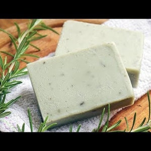 Könnte beinhalten: Zwei handgemachte Seifenstücke in hellgrüner Farbe mit Kräutern. Die Seife liegt auf einem weißen Handtuch und einer Holzoberfläche. Neben der Seife liegt ein Rosmarinzweig.