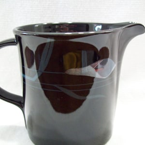May include: A glossy black ceramic pitcher featuring a curved handle and a pouring spout. The exterior surface is decorated with thin, abstract horizontal lines in muted blue and white tones that wrap around the body of the vessel.
