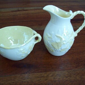 May include: A cream-colored ceramic creamer and sugar bowl set featuring a glossy finish and embossed floral details. The creamer has a twisted handle and a pouring spout, while the sugar bowl includes two side handles and a wide opening.