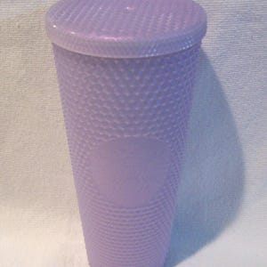 May include: A lavender-colored tumbler with a textured, studded surface and a matching lid. The cup has a cylindrical shape and a smooth, oval emblem on the front. The lid fits snugly on top, creating a secure seal. The cup is designed for cold beverages.