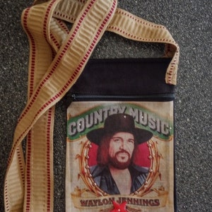 May include: A black fabric crossbody bag with a zipper closure. The front of the bag features a vintage-style illustration of Waylon Jennings with the text "Country Music" and "Waylon Jennings". A red star is attached to the bag.