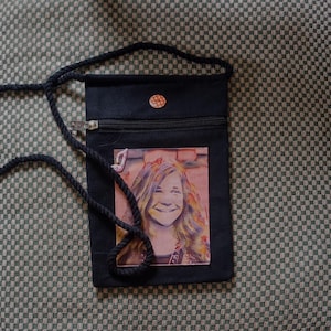 May include: Black canvas crossbody bag with a zipper closure and a small button. The bag features a colorful image of a woman with long hair.
