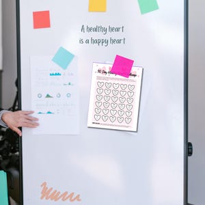 Puede incluir: Una pizarra blanca de borrado en seco con una nota adhesiva rosa, verde y amarilla. La pizarra tiene el texto "Un coraz&oacute;n sano es un coraz&oacute;n feliz" escrito en ella. Un desaf&iacute;o de 30 d&iacute;as para el coraz&oacute;n imprimible est&aacute; unido a la pizarra con una nota adhesiva rosa.