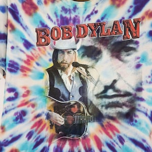 May include: Tie-dye t-shirt featuring a portrait of Bob Dylan with the text "BOB DYLAN" in a red and brown font. The shirt has a colorful tie-dye pattern in shades of blue, purple, pink, and white.