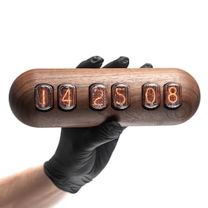 May include: A horizontal, oval-shaped wooden clock featuring six glowing orange nixie tubes arranged in a row. The tubes display the numbers 1, 4, 2, 5, 0, and 8, emitting a warm, amber light against the dark, polished wood grain of the casing.