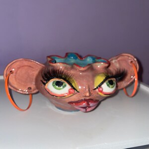 May include: A ceramic planter shaped like a face with large eyes, long eyelashes, and a pink and yellow painted design. The planter has orange hoop earrings.