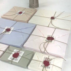 May include: A stack of cream, white, light blue, pink, and green envelopes with red wax seals. Each envelope is tied with twine and has a label that reads "Artesanos del Papel" in a decorative font.