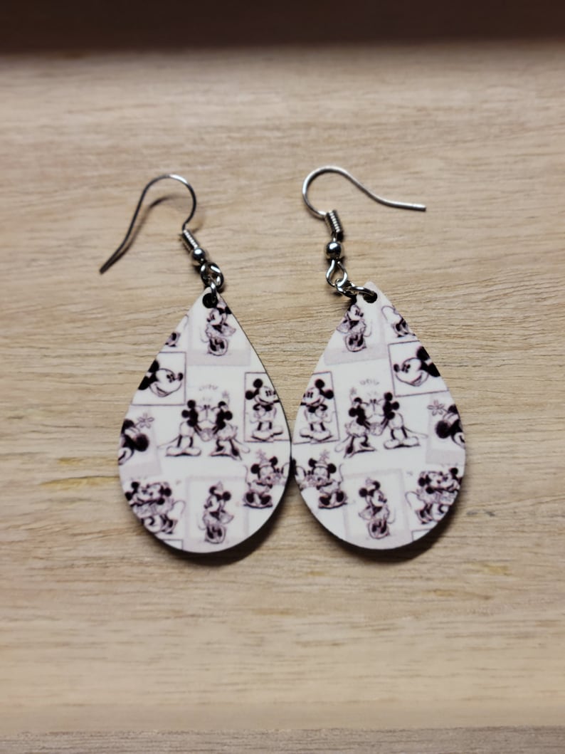 K&ouml;nnte beinhalten: Ein Paar Ohrringe mit Tropfenform. Die Ohrringe haben einen wei&szlig;en Hintergrund mit einem schwarz-wei&szlig;en Muster von Mickey Mouse und Minnie Mouse, die sich k&uuml;ssen.