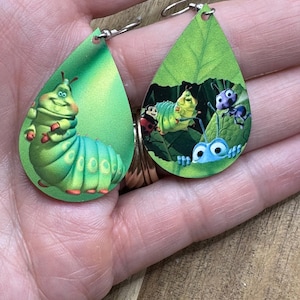 May include: Teardrop-shaped earrings featuring cartoon characters. One earring shows a green caterpillar with red accents. The other earring depicts a scene with a caterpillar, ladybug, and other insects on a green leaf. Silver-colored ear wires.