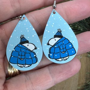 May include: Teardrop-shaped earrings featuring a cartoon character in a blue winter coat and hat, set against a light blue background with white snowflakes. The earrings have silver-colored hooks.