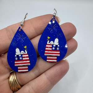 May include: Teardrop-shaped earrings with a blue background featuring Snoopy and Woodstock on a red, white, and blue design. The earrings have silver-colored hooks and depict fireworks.
