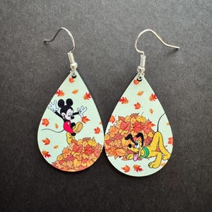 May include: A pair of teardrop-shaped dangle earrings featuring a light green background with a fall theme. One earring shows Mickey Mouse jumping into a pile of orange and red autumn leaves, while the other shows Pluto playing in the leaves.