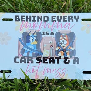 May include: A white license plate with the text "BEHIND EVERY momma IS A CAR SEAT & A hot mess." The plate features cartoon images of two dogs in car seats, with a floral pattern in the background.