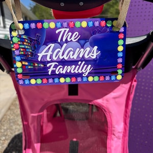 May include: A personalized license plate with the text "The Adams Family" in white script against a purple and blue background. The plate is trimmed with colorful, circular lights and is attached to a pink stroller.