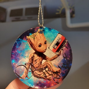 May include: A round, colorful, galaxy-themed car charm featuring a cartoon image of Groot from Guardians of the Galaxy holding a cassette tape. The text on the cassette tape reads "Awesome Mix Vol. 2".