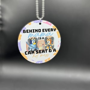 May include: A round white car charm with a colorful floral pattern. It features two cartoon dogs in car seats and the text "Behind every mama is a car seat & a hot mess".