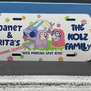 May include: A white license plate with a cartoon image of two dogs in blue and pink pajamas. The text on the plate reads "Janet & Rita's Driving School" and "The Kolz Family".