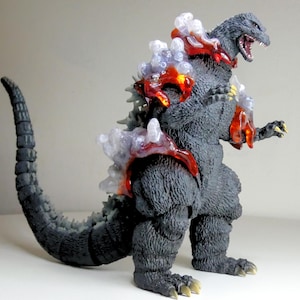 May include: A detailed figurine of Godzilla, a fictional monster. The figure is primarily dark gray with lighter gray dorsal fins. It features translucent red and white accents on its back and chest, with its mouth open, revealing teeth.