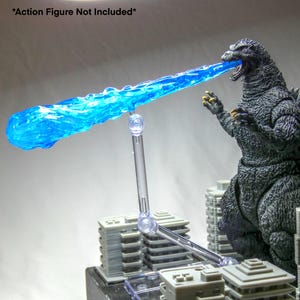 May include: A clear plastic stand supporting a translucent blue energy blast effect. The effect is positioned in front of a Godzilla action figure, with miniature building models in the foreground. The text "*Action Figure Not Included*" is in the top left corner.
