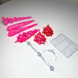 May include: A collection of translucent plastic action figure accessories. Includes pink energy blasts, red spiky orbs, a clear stand, and support pieces. These parts are designed to enhance the display of action figures.