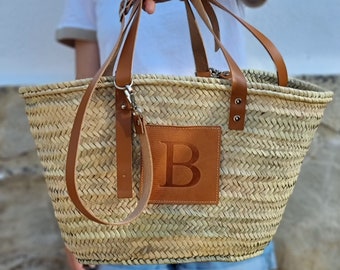 May include: Woven straw tote bag with brown leather handles and a leather patch with the letter "B". The bag has a rectangular shape and is held by a person. The bag is a natural color.