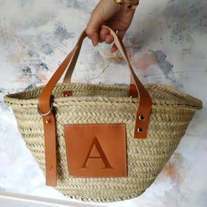 Puede incluir: Un bolso tote de paja natural tejido con asas de cuero marr&oacute;n y un parche de cuero marr&oacute;n con la letra "A" en relieve.