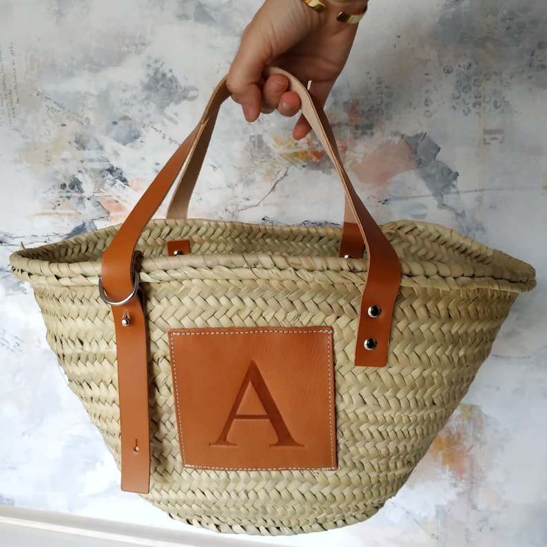 Puede incluir: Un bolso tote de paja natural tejido con asas de cuero marr&oacute;n y un parche de cuero marr&oacute;n con la letra "A" en relieve.