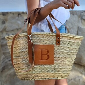 May include: Woven straw tote bag with brown leather accents. Features a brown leather patch with the letter "B" embossed on the front. Includes a shoulder strap and two handles.