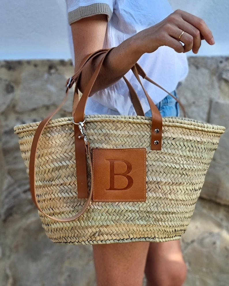 May include: Woven straw tote bag with brown leather accents. Features a brown leather patch with the letter "B" embossed on the front. Includes a shoulder strap and two handles.